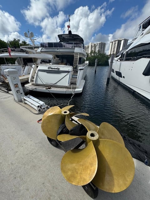 Brass boat propeller in foreground with luxury yachts docked at marina and blue sky with clouds
