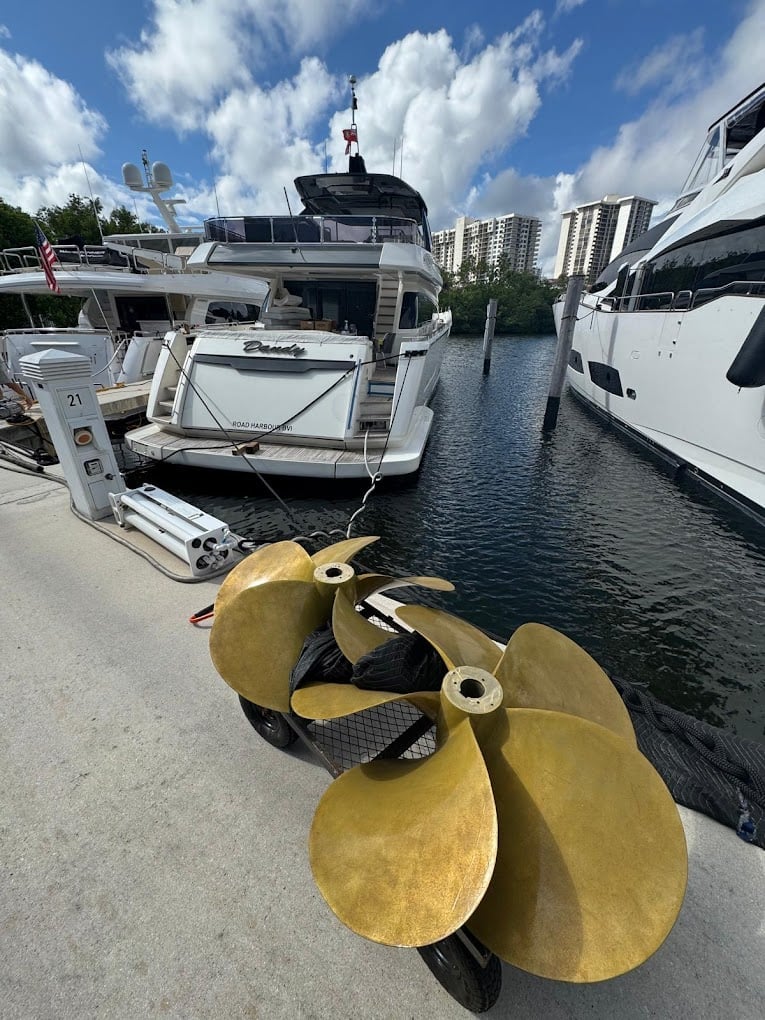Brass boat propeller in foreground with luxury yachts docked at marina and blue sky with clouds