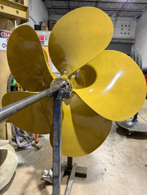 Large yellow four-bladed propeller mounted on a metal shaft in an industrial warehouse setting.