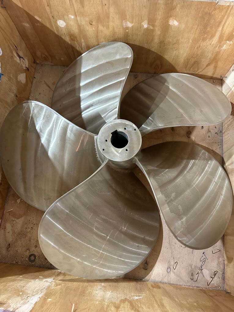 Four-bladed metal propeller with white center hub resting on wooden surface inside wooden crate