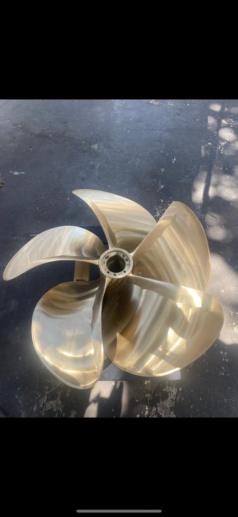Brass boat propeller with four curved blades displayed on a dark wet surface, casting shadows in natural light