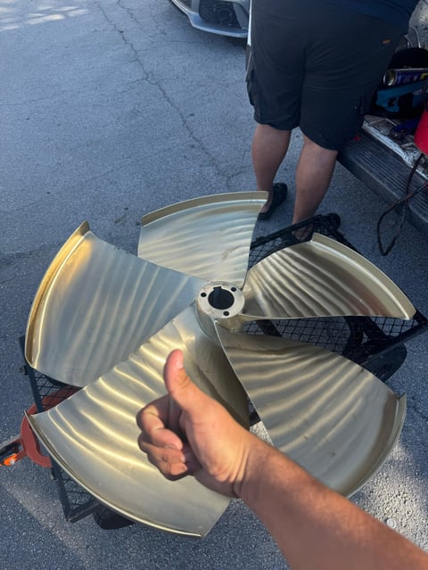 Person holding a large three-blade boat propeller on asphalt with another person visible in background.
