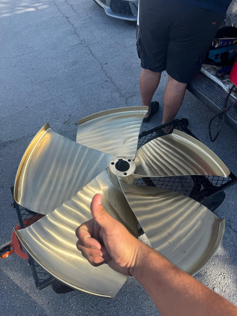 Person holding a large three-blade boat propeller on asphalt with another person visible in background.